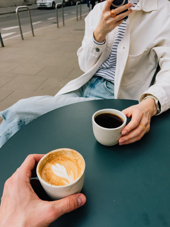 First person view: two friends are sitting at table outside cafe, enjoying coffee, capturing moment and sharing pleasant experience together. Friends relaxing at outdoor cafe, sipping coffeeの写真素材
