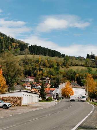 Two delivery vans driving along winding road toward small village nestled in Black Forest mountains of Germany on sunny autumn day with vibrant foliage. Delivery vans navigating curving mountain roadの写真素材