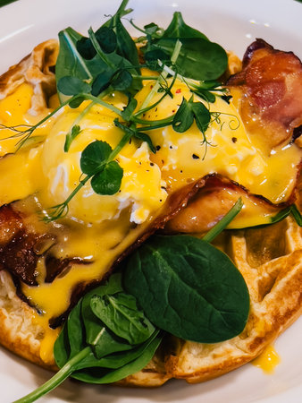 Close up of savory waffle topped with poached eggs, crispy bacon, creamy hollandaise, fresh spinach, creating appetizing brunch dish. Gourmet brunch plating. Waffle, eggsの写真素材