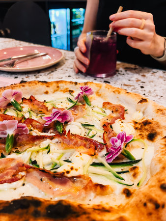 Woman is holding purple cocktail with straw and enjoying gourmet pizza topped with zucchini, bacon, edible flowers, and white sauce in restaurant setting. Woman sipping purple cocktail, enjoying pizzaの写真素材