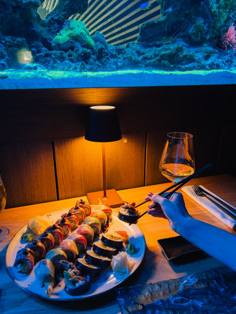 Woman using chopsticks picking up piece of sushi roll from plate full of colorful sushi, in restaurant with large illuminated aquarium. Woman eating sushi roll near large illuminated aquarium. Dateの写真素材