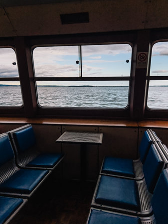 Empty passenger seats and small table inside ferry boat navigating on Lake with view on water and cloudy sky through large windows. Vacant ferry seats, small table, large windows revealing Lake watersの写真素材