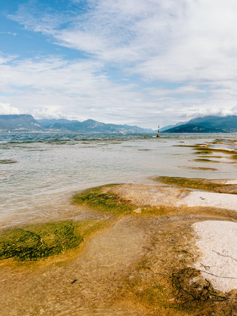 Water flowing over rocks and vibrant algae at Lake Garda on sunny, windy day, framed by majestic mountains and fluffy clouds in background, creates serene landscape. Cascading water, majestic mountainの写真素材