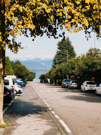 Lake and Alps mountains are visible at end of quiet road bordered by parked cars and trees with yellow autumn leaves in sunny day. Alpine peaks rising in sunlit background. Naturaの写真素材