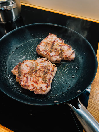 Two seasoned pork neck steaks are frying in hot pan on modern induction stove, creating delicious aroma and steam, classic culinary scene. Sizzling pork neck steaks browning in cast iron skilletの写真素材