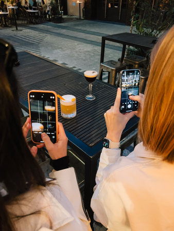 Two women are taking photos of cocktails with their smartphones, sitting at table in modern bar, capturing moment and drinks. Women photographing colorful cocktails Trendy beverage experienceの写真素材