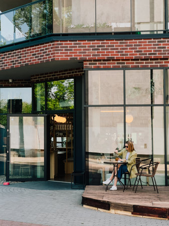Young woman enjoying break while pouring tea at table outside modern cafe featuring glass and brick...