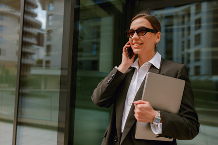 Businesswoman smiling and talking on phone, holding a laptop outside modern office buildingの写真素材