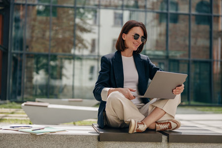 A stylish professional businesswoman is working on her laptop outdoors in a modern urban settingの写真素材