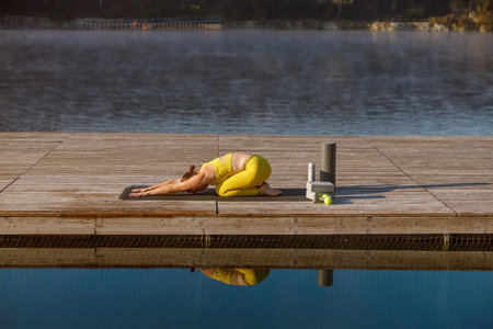 A Woman Practicing Yoga by the Serene Lake at Sunrise in a Peaceful Natural Settingの写真素材