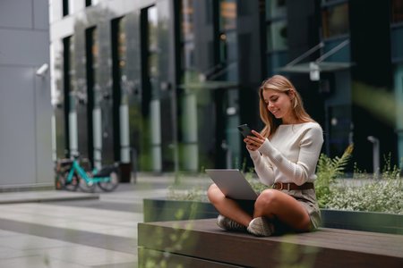 A young woman is working outdoors, utilizing her laptop and phone effectively while enjoying nature