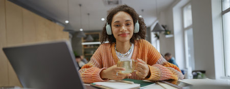 A woman at a table with laptop and headphones, enjoying coffee and smiling