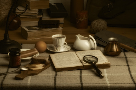 An open book, a cup of coffee, a magnifier and food on the table. A bag, books, a candle and other things on the windowsillの写真素材