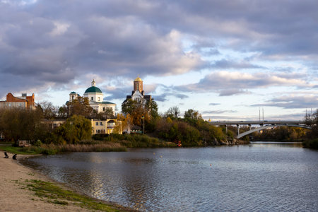 An evening view reveals a church on the riverbank under autumns cloudy yet sunny sky. The golden...