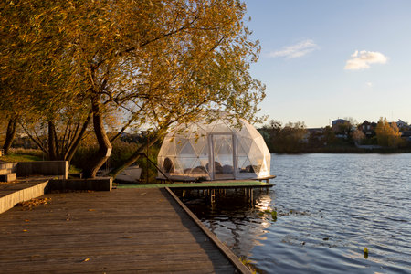 A transparent dome structure stands on the river bank on a sunny autumn day, surrounded by golden trees and calm water. Inside, soft light and warmth create a peaceful space for rest, reflection, and quiet relaxation.の写真素材