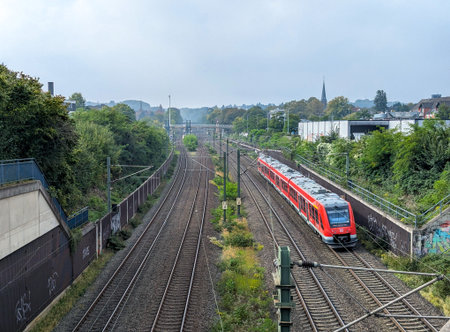 Cologne, Germany - September 30, 2025: A commuter train passes through Frechen-Konigsdorf station, with railway tracks stretching into the distance under a calm sky and urban surroundings.の写真素材