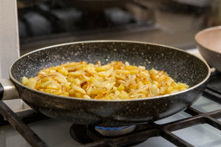 Potato slices with onions are fried in a pan until golden and crispy, with appetizing texture and warm kitchen light.の写真素材