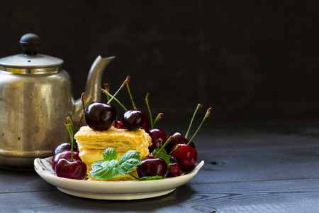 Still life with a teapot, a piece of puff cake, a sprig of mint and cherries. Dark background. Food photo.の写真素材