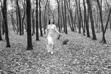 portrait of young red-haired woman in autumn forest. girl in a long white dress with basket. Beautiful women's body in silk nightwear. nymph.wreath of flowersの写真素材