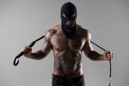 Muscular shirtless young man with whip in a mask. Men tattoo casual fashion. Portrait of a handsome male model. Muscular athletic sexy male witの写真素材