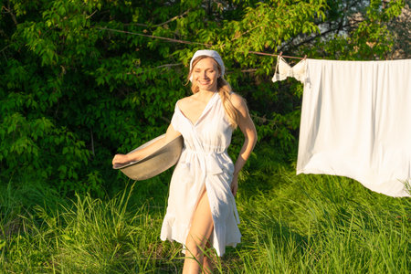 Beautiful young woman in white dress and hat drying clothes on clothesline in summer gardenの写真素材