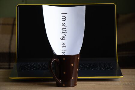 laptop on a table in a room at home and a sheet with the inscription in English sitting at home in the quarantine period in a cup next toの写真素材