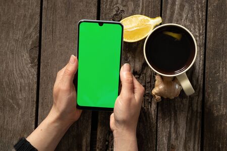 female hands hold a mobile phone and a cup of tea with lemon on an wooden tableの写真素材