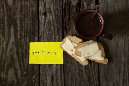 hot black coffee and white bread and butter on an wooden table and the words in English good morning on yellow paperの写真素材