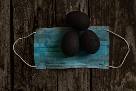 medical mask and painted black egg on Easter day on an old wooden table during the pandemicの写真素材
