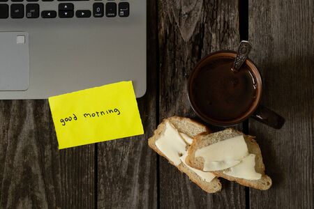 coffee and white bread and butter on an wooden table and the words in English good morning on yellow paper and a laptopの写真素材