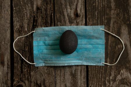 medical mask and painted black egg on Easter day on an old wooden table during the pandemicの写真素材