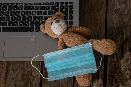 children's bear in a medical mask lies on a laptop on a wooden table at home in the period of quarantineの写真素材