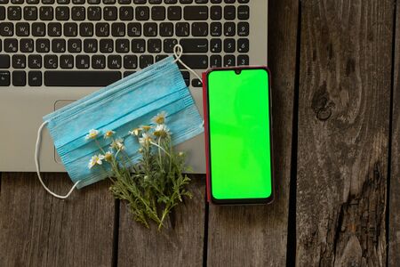 laptop medical mask and daisy flowers and a phone with a green screen on a tableの写真素材