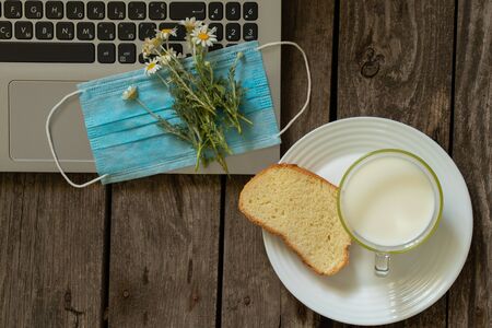 medical mask a piece of bread and a glass of milk on a table at home in the kitchen next to a laptopの写真素材