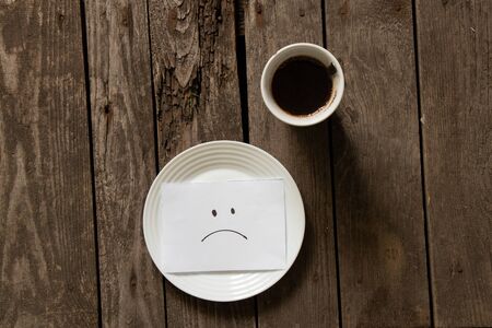empty plate with a sad smiley on paper on a wooden table and a cup of coffeeの写真素材