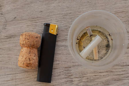 cigarette filter floats in a plastic cup and a lighter next to a cork from under the wine on the tableの写真素材