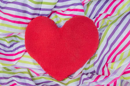 pillow in the shape of a red heart lies on a multi-colored fabric on the bedの写真素材