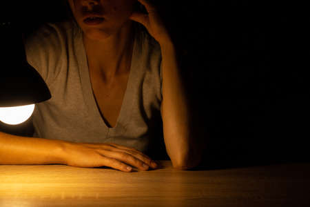 girl sitting at a table in the light of a table lamp in a dark roomの写真素材