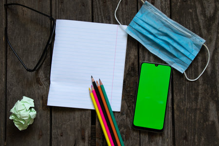 medical mask phone with a green screen next to an open notebook with pencils on the table, work in quarantine from homeの写真素材