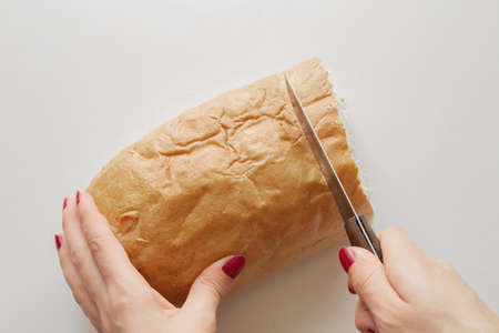 female hand cuts a piece of bread with a knife on a white table in the kitchen at home