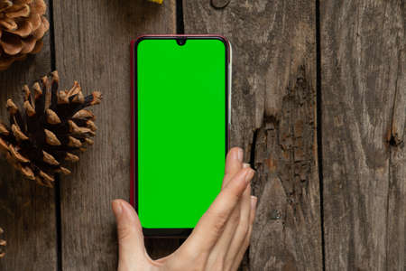 a phone with a green screen lies on a wooden table in the New Year's decor near pine conesの写真素材