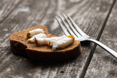 a piece of black bread with bacon and a fork on a wooden table for lunchの写真素材