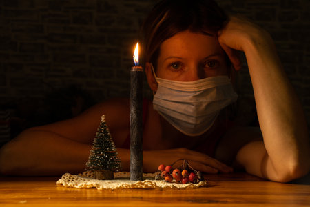 a girl in a medical mask sits carrying a burning candle and a small Christmas tree on the table in the dark, new year alone in quarantineの写真素材