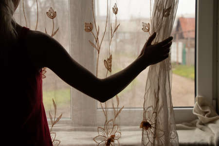 a woman's hand opens the curtain on the window in the bedroom, view from the window of the house to the streetの写真素材