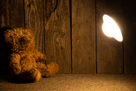 brown teddy bear sits on the floor in the dark by the light of a table lampの写真素材