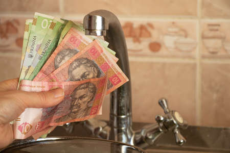 a woman's hand holds a Ukrainian 10 20 hryvnia near the kitchen tap, water prices