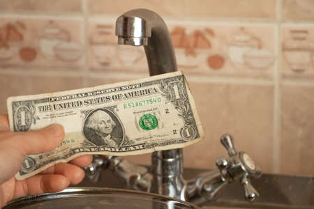 female hand holds one dollar near kitchen faucet, water prices