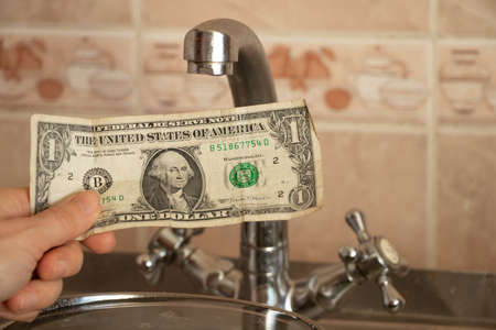 female hand holds one dollar near kitchen faucet, water pricesの写真素材