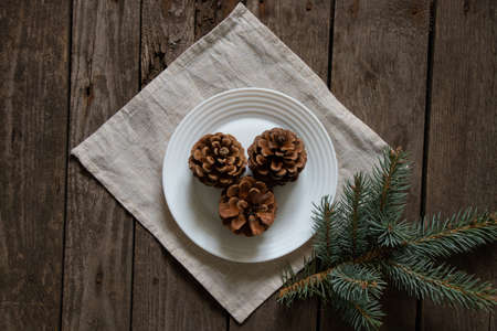 cones on a white plate next to a branch of a Christmas tree on a tablecloth on a wooden table, New Year's table decorの写真素材
