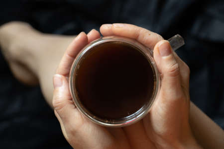 girl holding a hot cup of coffee with two hands on a black background close-upの写真素材